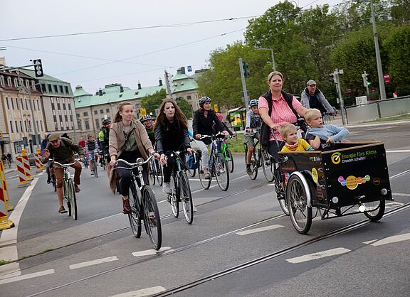 Gruppe von Radfahrenden auf einer Straße mit Straßenbahnschienen in Dresden. Im Vordergrund fährt eine Frau mit einem schwarzen Lastenrad, in dem zwei kleine Kinder sitzen. Neben ihr fahren weitere Personen mit verschiedenen Fahrrädern.